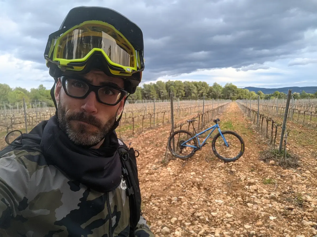 Ger after a vineyard ride — helmet and 100% goggles up, his muddy hardtail parked between the vines.